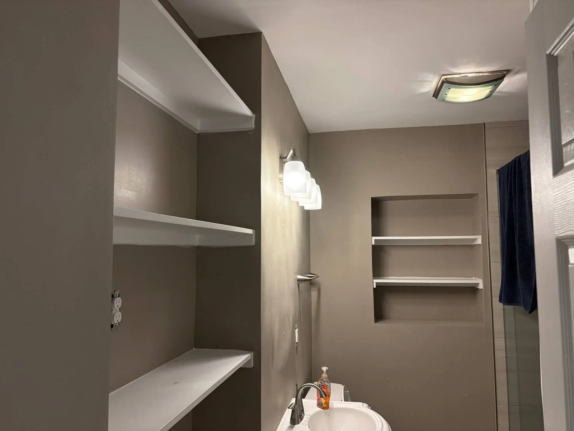 Elegant bathroom transformation by 1st Coast Painting featuring matte taupe walls contrasted with crisp white built-in shelving and modern vanity lighting.
