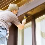 Professional painter applying stain to wood trim and siding on a house exterior, illustrating the labor involved in house painting cost.