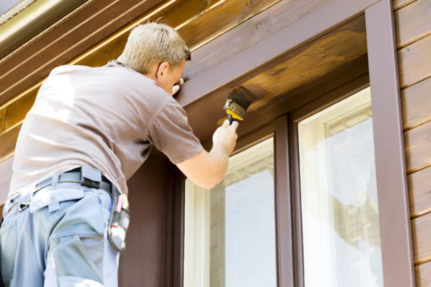 Professional painter applying stain to wood trim and siding on a house exterior, illustrating the labor involved in house painting cost.
