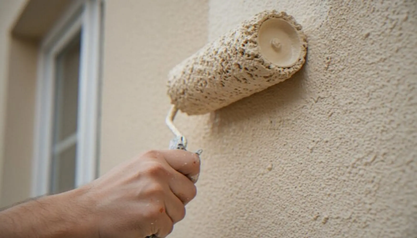 Roller applying light exterior paint to a stucco wall in mild, dry weather, illustrating the best time of year for exterior house painting.