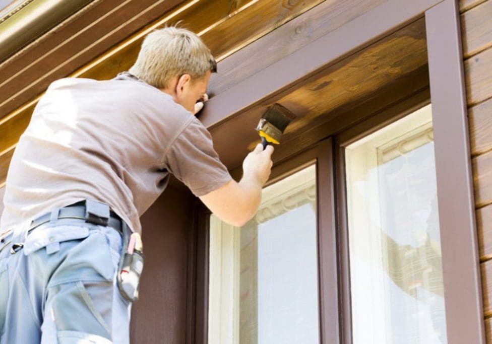 Professional painter applying stain to wood trim and siding on a house exterior, illustrating the labor involved in house painting cost.